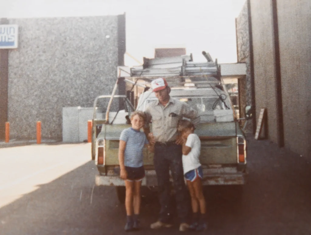 A man stands between two children in front of an old pickup truck with ladders on top, parked in an alley between two buildings on a sunny day. The man has his arms around the children.