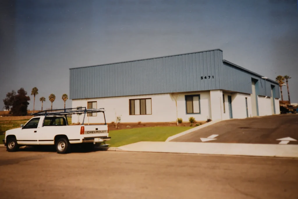 A white pickup truck is parked in front of a one-story industrial building with blue siding and the number 5471. The building has several windows and a well-kept lawn, with palm trees visible in the background.