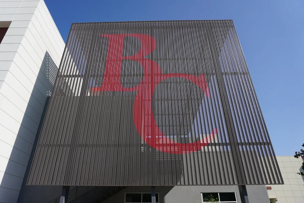 A large metal screen with the overlapping red letters B and C prominently displayed, set against a blue sky and part of a modern building.