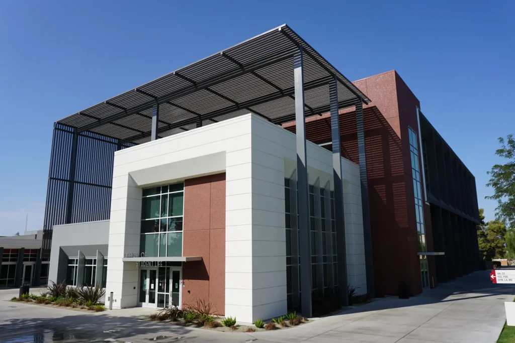 A modern building with white and red-brown walls, large windows, and a prominent metal canopy structure on the roof, set against a clear blue sky.