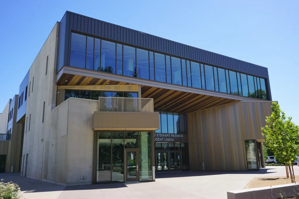 A modern building with large glass windows and wood-paneled exterior, featuring an overhanging upper floor and a shaded entrance under clear blue sky.