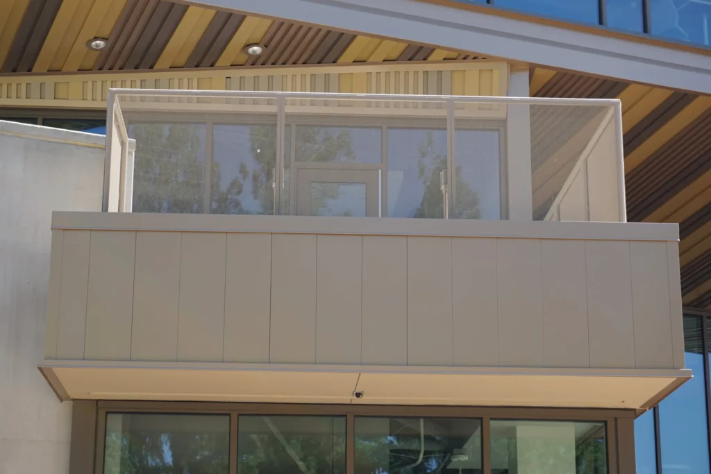 A modern building featuring a balcony with frosted glass panels and beige metal framing, reflecting trees and the sky in its windows beneath an angled roof with wooden soffits.
