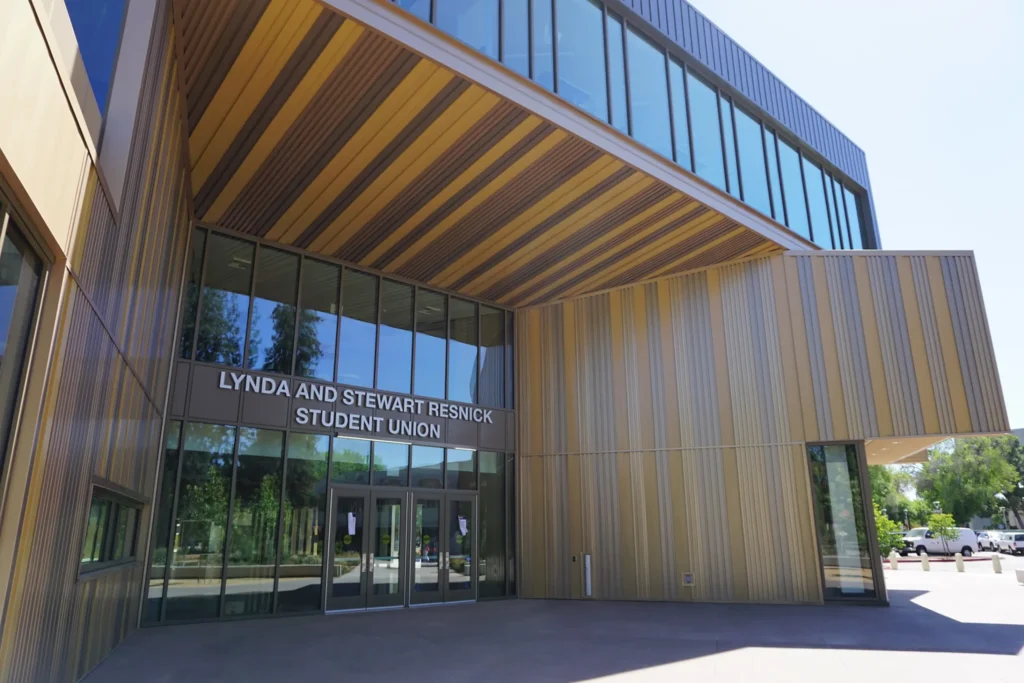Modern building with large windows and a wooden exterior. Above the glass entrance doors, a sign reads Lynda and Stewart Resnick Student Union. Trees and bright sunlight are reflected in the glass.