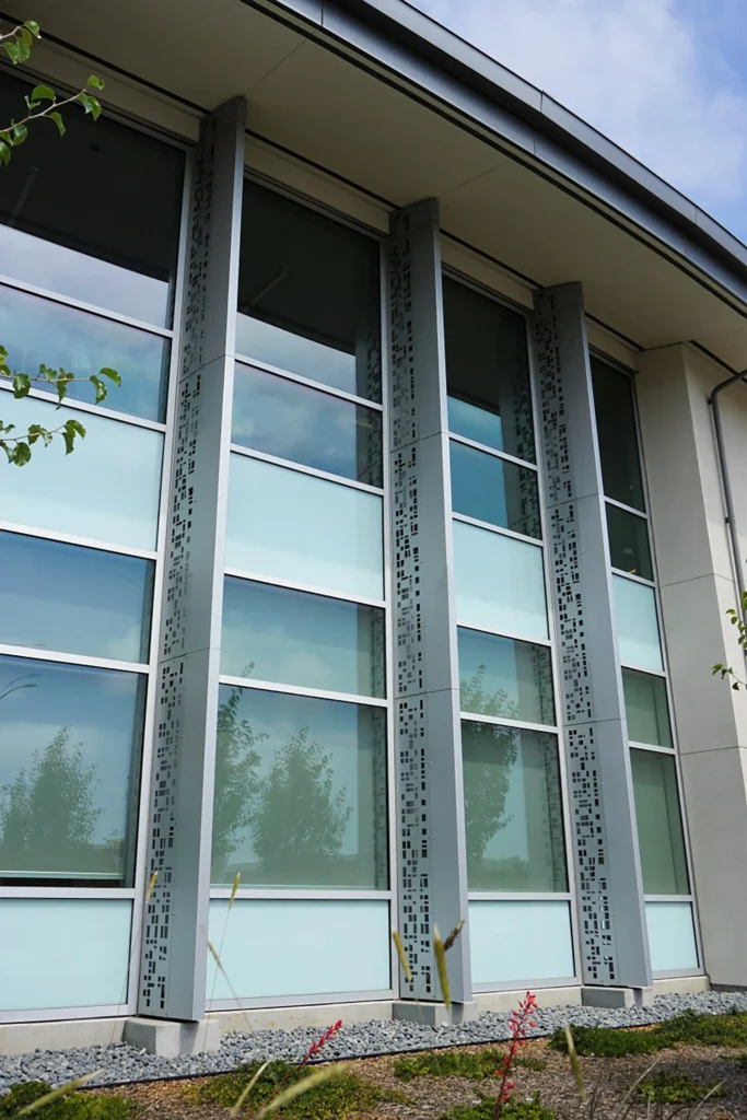 A modern building facade with large glass windows, vertical metal panels with perforated patterns, and a landscaped area with small plants in the foreground. The sky is partly cloudy.