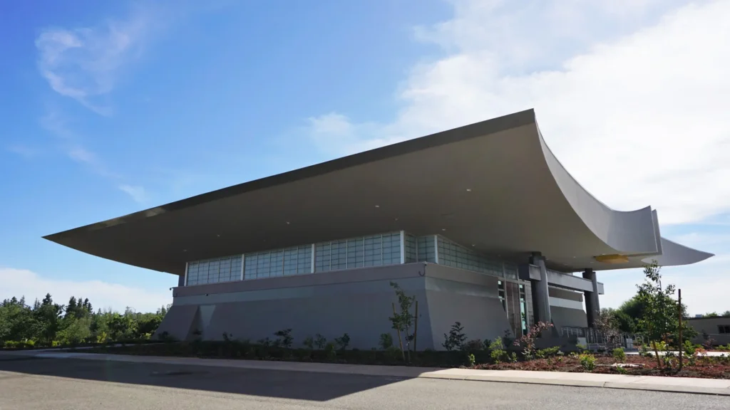 A modern building with a large, curved overhanging roof, glass block windows, and landscaped greenery in front, photographed under a bright blue sky.