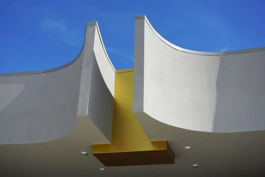 Modern building facade with two white, curved vertical structures separated by a yellow section, seen from below against a clear blue sky. Four recessed lights are visible under the overhang.