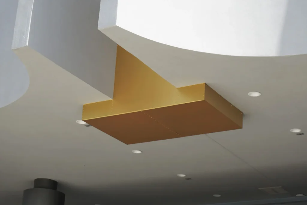 A gold, T-shaped architectural feature is mounted on a white ceiling with recessed lights, featuring modern, geometric shapes and shadows.