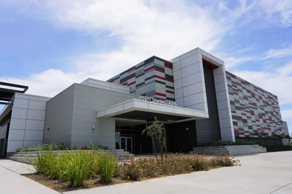 A modern building labeled Performing Arts with geometric gray, white, and red panels, large windows, and landscaping with green plants and flowers under a partly cloudy sky.