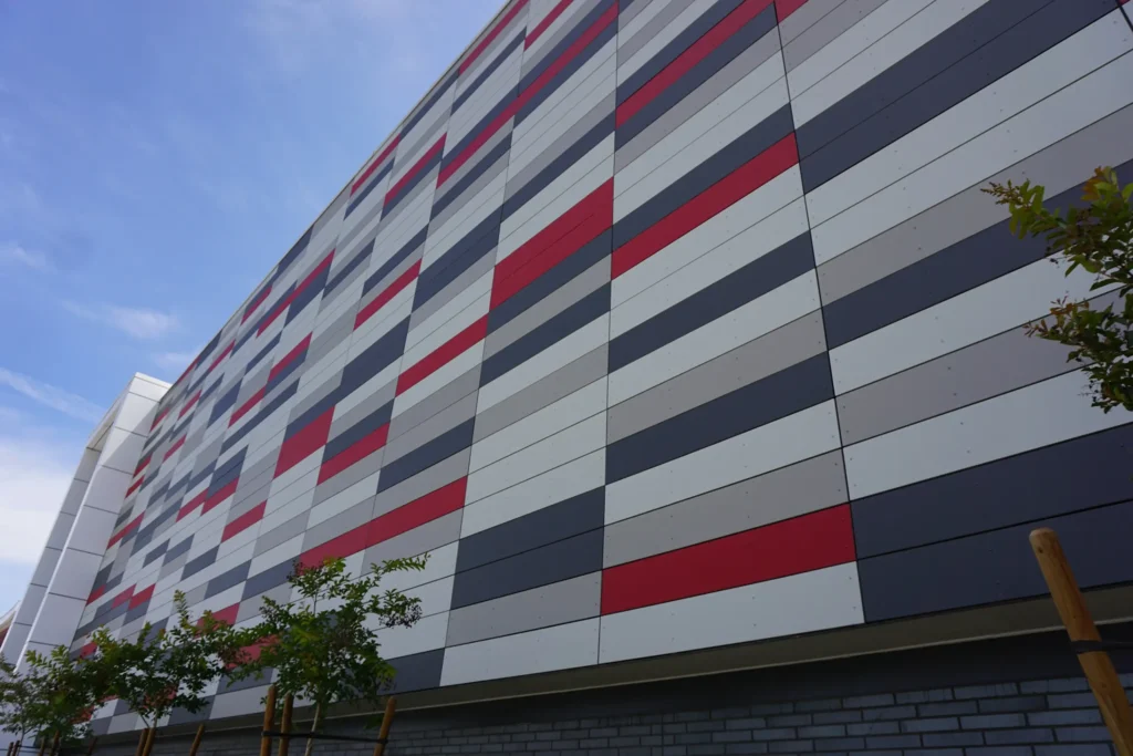 A modern building facade with a geometric pattern of red, gray, black, and white horizontal panels, viewed from below. A few small trees are visible near the base against a blue sky.