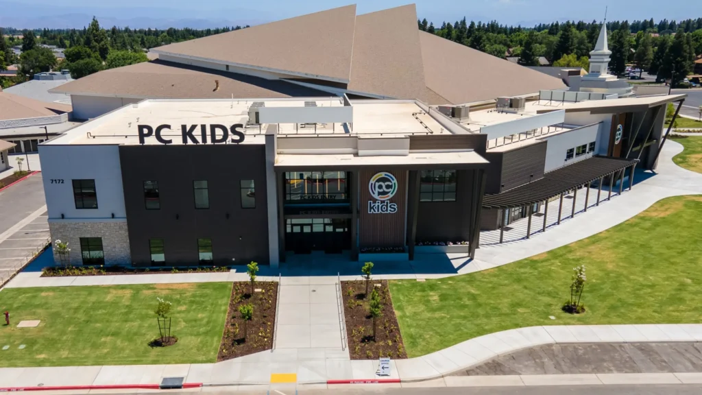 A large modern building labeled PC KIDS with a neatly landscaped lawn, wide sidewalks, and a circular driveway in front, under a clear sky.