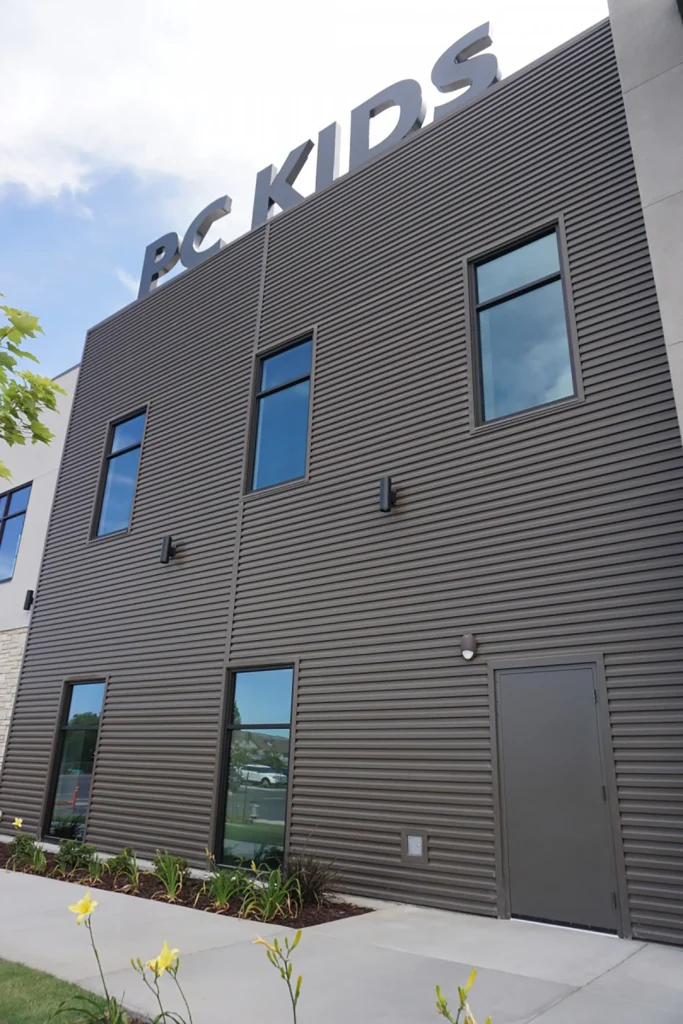 A modern building with a ribbed brown facade has large windows, a gray door, and a sign on the roof reading PC KIDS. Yellow flowers and a sidewalk are in the foreground.
