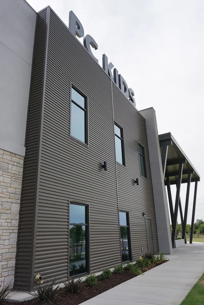 Exterior view of a modern building with a sign reading PC KIDS on the roof. The structure features large windows, vertical pillars, and a mix of gray siding and stone accents, with a sidewalk and landscaping in front.