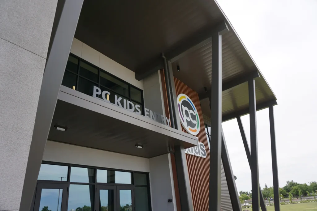 Modern building exterior with large windows and a sign that reads “PC KIDS ENTRY” above the entrance. The structure features tall support beams and a partial view of a colorful circular logo.
