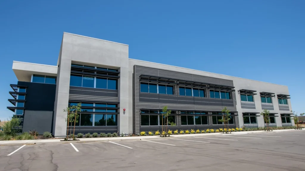 A modern two-story commercial office building with large windows and a gray facade sits in front of an empty parking lot under a clear blue sky.