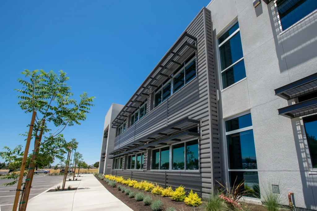 Modern commercial building with large windows, metal awnings, and a neat landscaped area featuring young trees and shrubs, under a clear blue sky.
