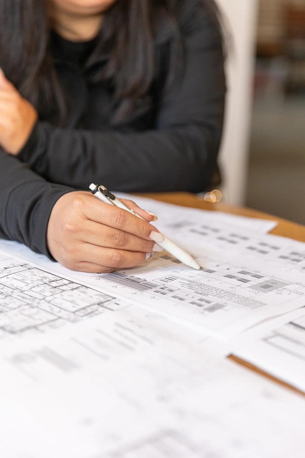 A person in a black long-sleeve shirt holds a pen and points to architectural blueprints on a table, with other plans and documents spread around.