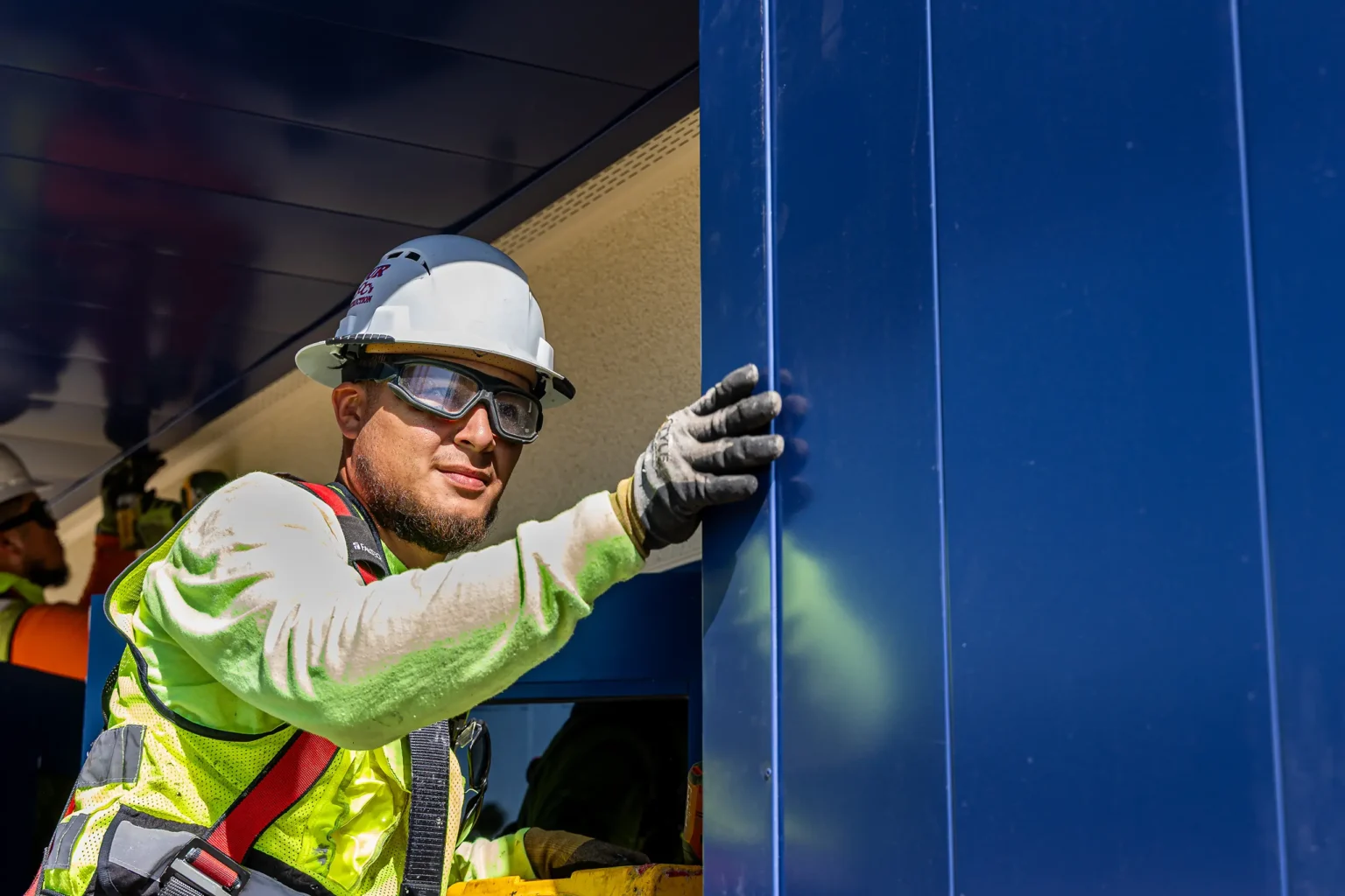 A construction worker wearing a hard hat, safety glasses, gloves, and a reflective vest is installing or inspecting a large blue panel on a building site, with sunlight illuminating his face.